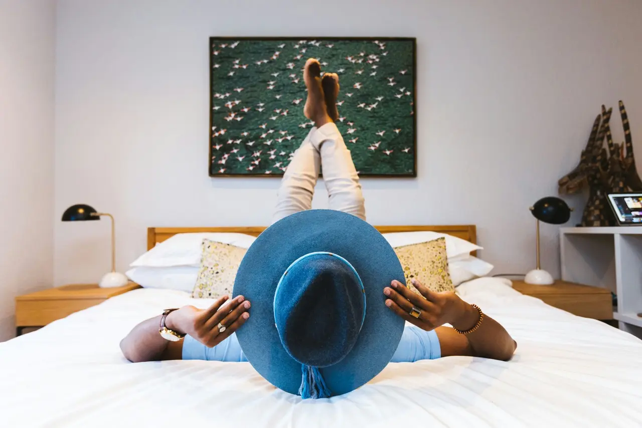 Pessoa deitada na cama de um quarto claro, com um chapéu azul sobre o rosto
